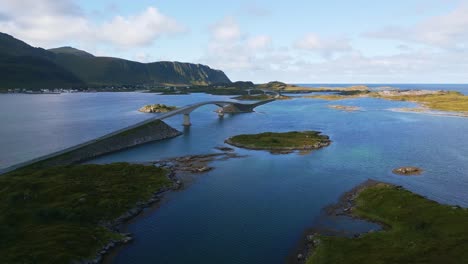 Flying-along-Fredvang-bridge-in-Lofoten-Northern-Norway-with-the-sea-ocean-water-shimmering-in-light-turquoise-blue-and-green-tones-on-a-sunny-day-in-summer-with-little-islands-surrounding-the-road