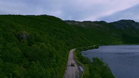 Following-a-golden-car-through-the-lush-green-natural-forests-of-northern-Norway-in-Lofoten-with-blue-lake-to-the-right-and-mountains-to-the-left-and-the-horizon