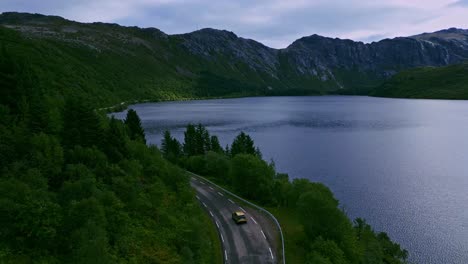 Aerial-shot-following-a-golden-car-through-the-green-forest-tree-landscape-in-Lofoten-Northern-Norway-towards-Nusfjord-with-the-road-winding-through-lakes-with-dramatic-mountains-in-the-background