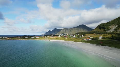 Flying-along-paradise-beach-in-Lofoten-Norway-with-view-towards-the-high-mountain-peaks-with-waves-crashing-along-the-white-sand-beach-shores-with-red-northern-houses-along-the-coast