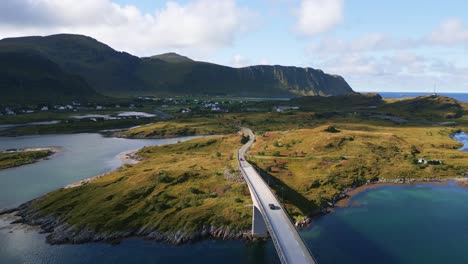 Following-the-black-car-driving-along-the-Fredvang-bridge-in-Lofoten-Northern-Norway-with-the-light-turquoise-blue-sea-ocean-water-towards-the-mountains-on-the-horizon-and-green-yellow-grass-below