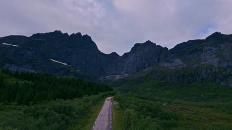Downwards-flying-shot-following-a-golden-car-through-the-green-grass-forest-tree-landscape-in-Lofoten-Northern-Norway-Europe-with-the-road-towards-the-horizon-of-lakes-with-mountains-in-the-background