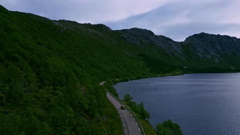 Aerial-shot-following-a-golden-car-through-the-green-forest-tree-landscape-in-Lofoten-Northern-Norway-with-the-road-winding-towards-Nusfjord-and-mountains-and-the-blue-lake-landscape-visible-in-summer