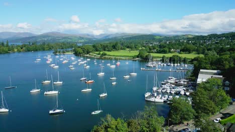 Descending-Aerial-Drone-View-Over-Boat-Moorings-And-Yachts-On-Lake-Windermere-At-Ferry-Nab-With-Colourful-Trees-Mountains-And-Blue-Sky-With-White-Clouds-On-Sunny-Summer-Morning