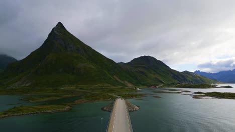 Flying-along-the-narrow-street-Fredvang-bridge-in-Lofoten-Northern-Norway-with-the-sea-ocean-water-shimmering-in-light-turquoise-blue-and-green-tones-in-summer-with-the-red-northern-houses-visible