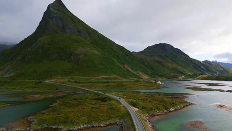 Flying-along-the-Fredvang-bridge-in-Lofoten-Northern-Norway-with-the-sea-ocean-water-shimmering-in-light-turquoise-blue-and-green-tones-on-the-steep-mountain-peek-covered-in-grass