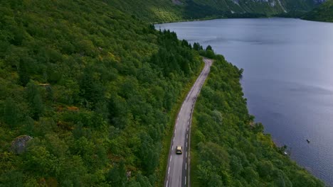 Downwards-facing-shot-of-golden-car-driving-along-the-scenic-road-on-the-coast-through-the-green-forests-in-Northern-Europe-Norway-Lofoten-with-view-towards-the-blue-ocean-and-high-dramatic-mountains