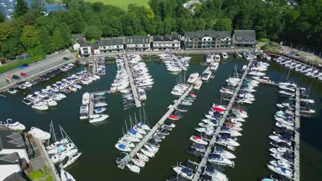 Forward-Moving-Aerial-Drone-View-Over-Boat-Moorings-And-Yachts-On-Lake-Windermere-At-Bowness-Marina-With-Colourful-Trees-On-Sunny-Summer-Morning-With-Visible-Algae-In-The-Lake