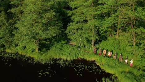 Drone-approaching-group-of-medieval-soldiers-walking-in-line-through-the-forest-on-the-shores-of-a-lake