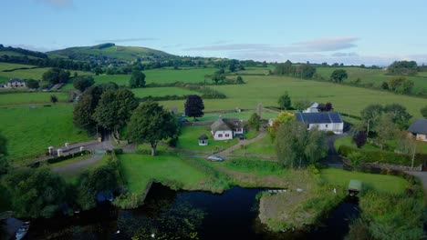 Picturesque-colorful-green-grass-and-trees-in-the-Irish-countryside-on-the-side-of-a-lough-with-a-traditional-cottage-on-the-shore-line,-drone-fly-over-on-a-sunny-bright-day