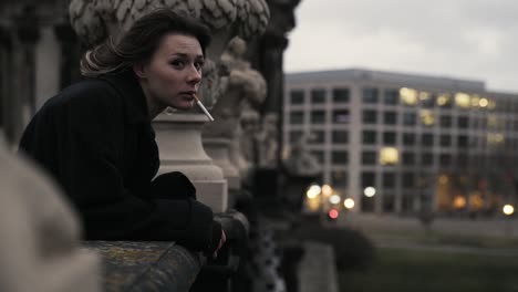 Young-woman-in-a-black-coat-standing-on-a-terrace-enjoying-the-view-and-smoke