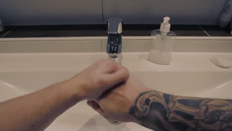 First-person-perspective-shot-of-a-young-man-with-tattoos-on-his-arm-washing-his-hands-with-soap-unser-running-water-in-a-modern-bathroom