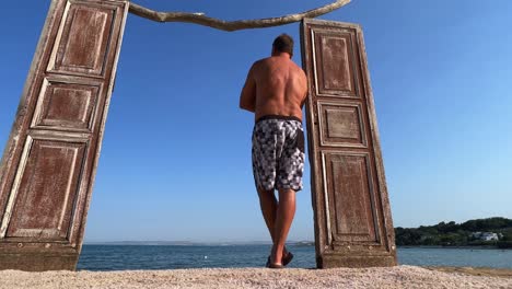 Decorative-seafront-wooden-door-and-back-view-of-man-standing-in-swimwear-leaning-against-doorframe