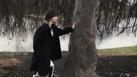 Young-woman-in-a-black-coat-standing-under-a-windy-tree-in-a-park-in-autumn-in-afternoon-4