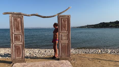 Unusual-footage-of-wooden-door-on-beach-for-shooting-set-facing-sea-with-man-in-swimwear-leaning-against-doorframe