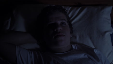 A-young-high-school-aged-teenager-lays-in-his-bed-at-night-looking-sad-and-serious