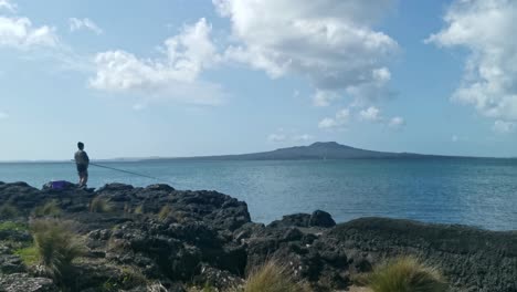 Singular-fisherman-at-Thorne-bay,-New-Zealand-with-magnificent-Rangitoto-volcano-in-the-background