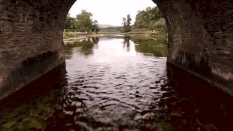 Fliegen-über-Einen-Fließenden-Fluss-Und-Fliegen-Unter-Einer-Alten-Backsteinbrücke-In-Der-Irischen-Landschaft