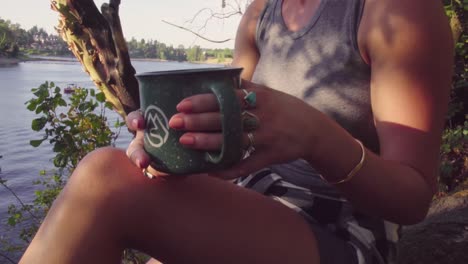 Cute-hipster-girl-relaxing-with-a-drink-in-a-hip-mug-next-to-a-river-or-lake
