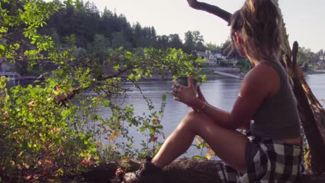 Cute-hipster-girl-sitting-by-a-lake-with-a-mug-to-drink-and-relax