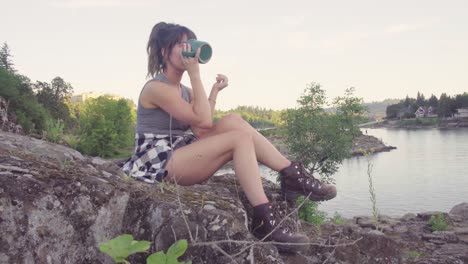 Cute-hipster-girl-drinking-from-a-mug-in-front-of-the-water-in-a-park
