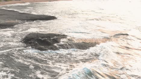 Drone-slow-motion-footage-of-rock-in-the-sea-close-to-the-seashore,-waves-wash-over-the-cliff