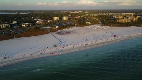Aerial-drone-shot-pulling-away-from-white-sandy-beach-with-drum-circle-in-Siesta-Key,-Florida-at-sunset