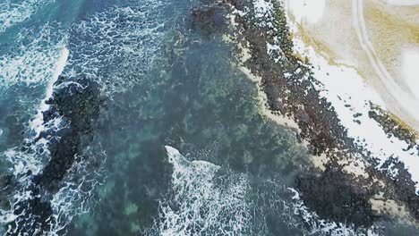 Aerial-shot-pointed-down-at-Sandgerdi,-Iceland-coast-showing-crashing-white-peaked-waves-and-clear-water-with-black-rocks