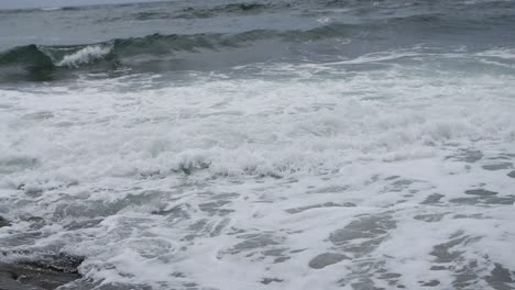 Turbulente-Schaumige-Wellen-In-Zeitlupe,-Die-Ihr-Lebensende-Am-Strand-Erreichen,-Erschossen-Vom-Strandfelsen