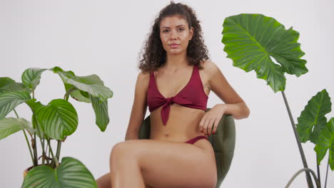 Dark-curly-haired-and-tanned-model-posing-on-a-chair-with-a-red-bikini-suit,-surrounded-by-plants,-shot-in-a-photo-studio