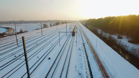 Drone-flying-backwards,-capturing-a-train-slowly-approaching-a-staition-during-the-winter,-surrounded-by-fresh-fallen-snow,-near-a-bicycle-road