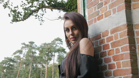 East-European-brunette-mysteriously-looking-into-the-camera,-surrounded-by-trees-and-a-brick-wall,-slow-motion-footage