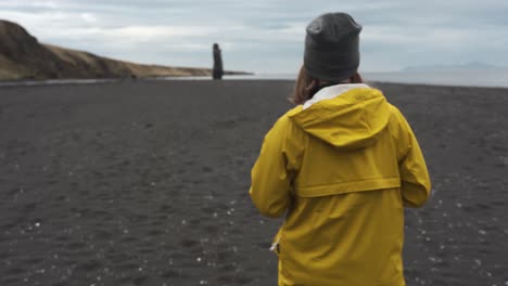 Woman-with-a-yellow-raincoat-walking-on-a-black-sand-beach-in-hvitserkur,-Vatnsnesvegur-located-in-Iceland