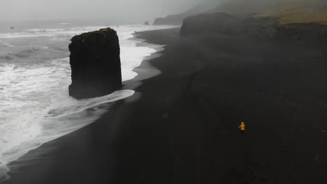 Girl-in-yellow-raincoat-walking-in-a-storm-along-a-black-beach-in-djupavogshreppur,-Iceland