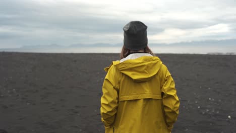 Woman-with-a-yellow-raincoat-walking-on-a-black-sand-beach-in-hvitserkur,-Vatnsnesvegur-located-in-Iceland-1