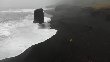 Girl-in-yellow-raincoat-walking-in-a-storm-along-a-black-beach-in-djupavogshreppur,-Iceland-1