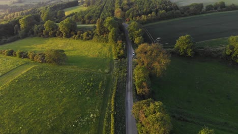 Aerial-cinematic-shot-of-english-country-road-in-green-countryside-during-sunset-with-bright-orange-glow