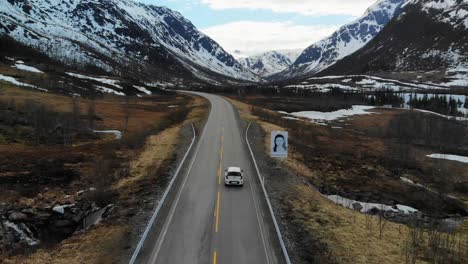 Picturesque-aerial-of-a-car-riding-on-a-road-between-forests-and-snowy-mountains