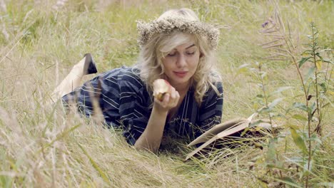 Attractive-blonde-woman-lies-in-a-field-of-grass,-eating-an-apple-and-reading-a-book