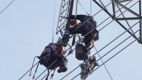 Two-utility-workers-in-hardhats-sit-on-electrical-high-voltage-power-lines-for-repair