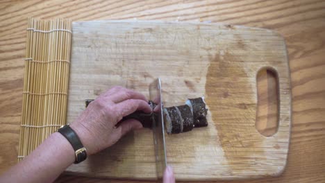 Older-Woman-Hands-Cutting-Sushi-Pieces-With-Knife-On-Cutting-Board-From-Top-View