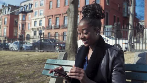 Attractive-Young-African-American-Woman-Using-Smartphone-Sitting-On-Park-Bench,-Receiving-Text-Message-That-Makes-Her-Laugh-And-Smile,-Sunny-Day-In-The-City