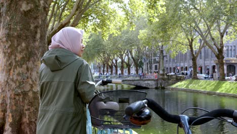 Muslimische-Frau-Neben-Altem-Fahrrad,-Das-Blick-Auf-Den-Kanal-In-Der-Düsseldorfer-Stadt-Genießt---Cam-B