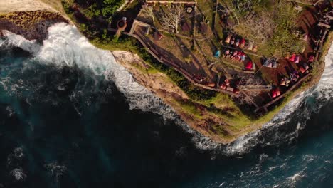 Birds-eye-drone-shot-of-cliff-side-beach-bar-with-waves-crashing-below-at-sunset-in-Bali