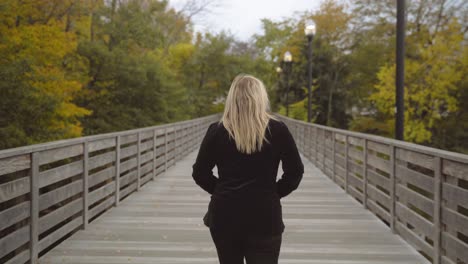 Blonde-hair-Caucasian-woman-walking-on-wooden-footbridge-exploring-alone,-follow-shot-from-behind-in-4k