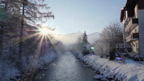 Sun-shining-bright-behind-snow-laden-mountains-during-sunrise,-and-water-stream-flowing-calmly-amidst-them