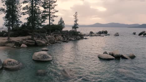 A-man-standing-in-the-rocks-of-Lake-Tahoe-beautiful-which-is-a-large-freshwater-lake-in-the-Sierra-Nevada-of-the-United-States-covered-with-tall-pine-trees,-aerial-clip