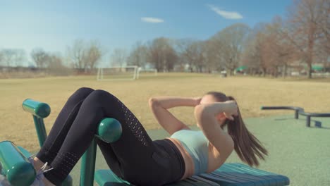 Young-Caucasian-Fit-Girl-Exercising-Doing-Sit-Ups-At-Outdoor-Gym-On-Sunny-Day,Staying-Focused-And-Determined,-Wearing-Sports-Bra-And-Yoga-Pants