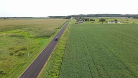 Drone-following-a-car-driving-along-a-rural-road-in-the-middle-of-farmer's-fields-with-various-crops,-aerial-view