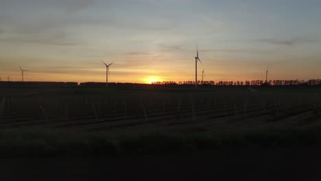 Wind-turbines-in-between-farming-fields-during-a-sunset,-travel-drone-shot